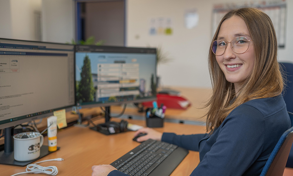 Person sitzt am Schreibtisch vor 2 Bildschirmen und lächelt in die Kamera
